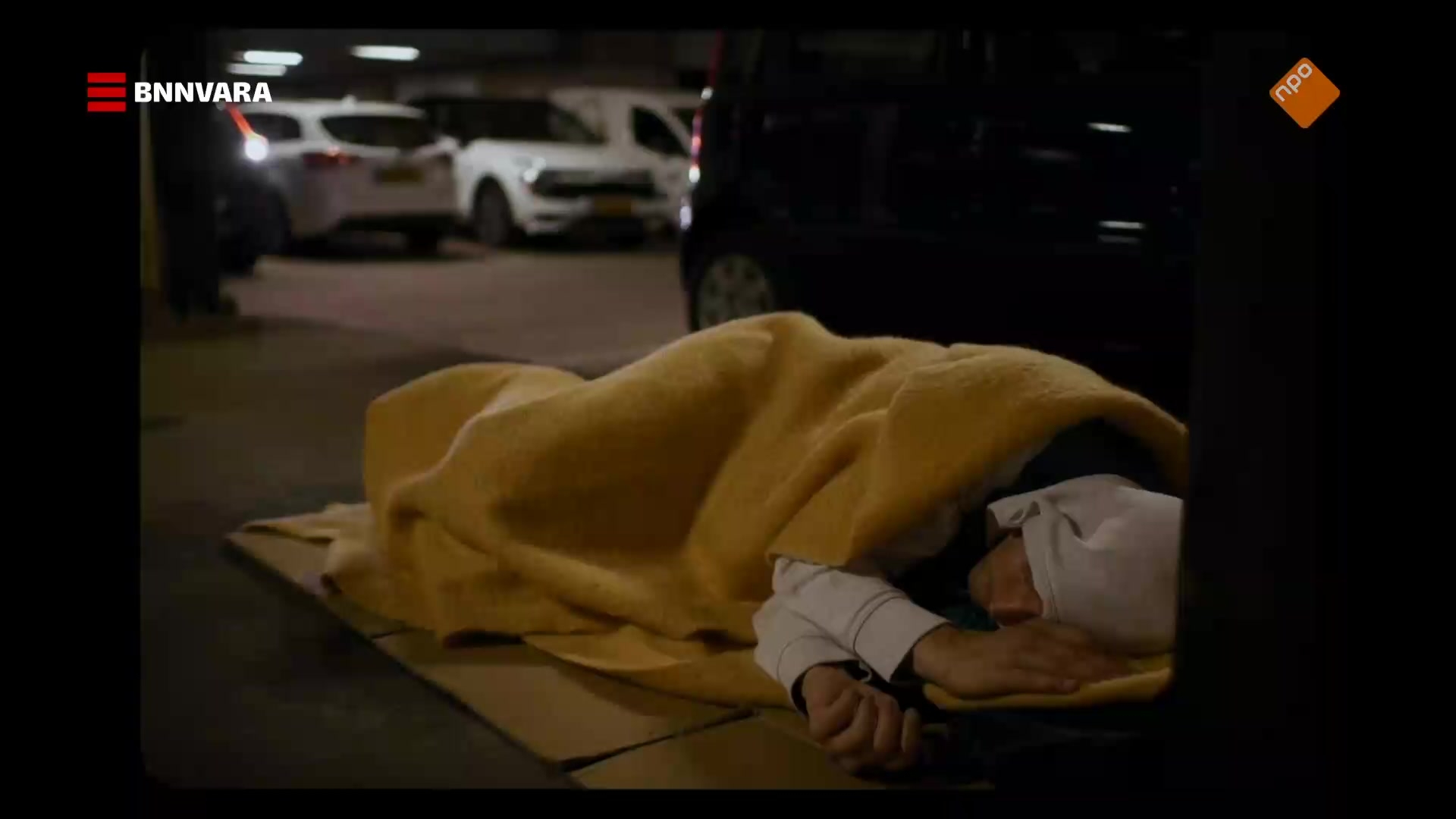 Persoon slaapt op straat, terwijl achter hem een auto wordt geparkeerd